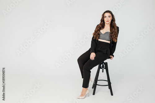 young girl with dark curly hair in a suit sits on a high chair on a white background, copy space