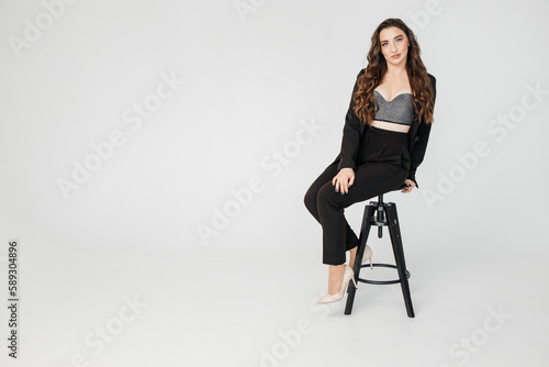 young girl with dark curly hair in a suit sits on a high chair on a white background, copy space