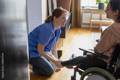 Happy nurse assisting woman with paraplegia while wearing shoes at home photo