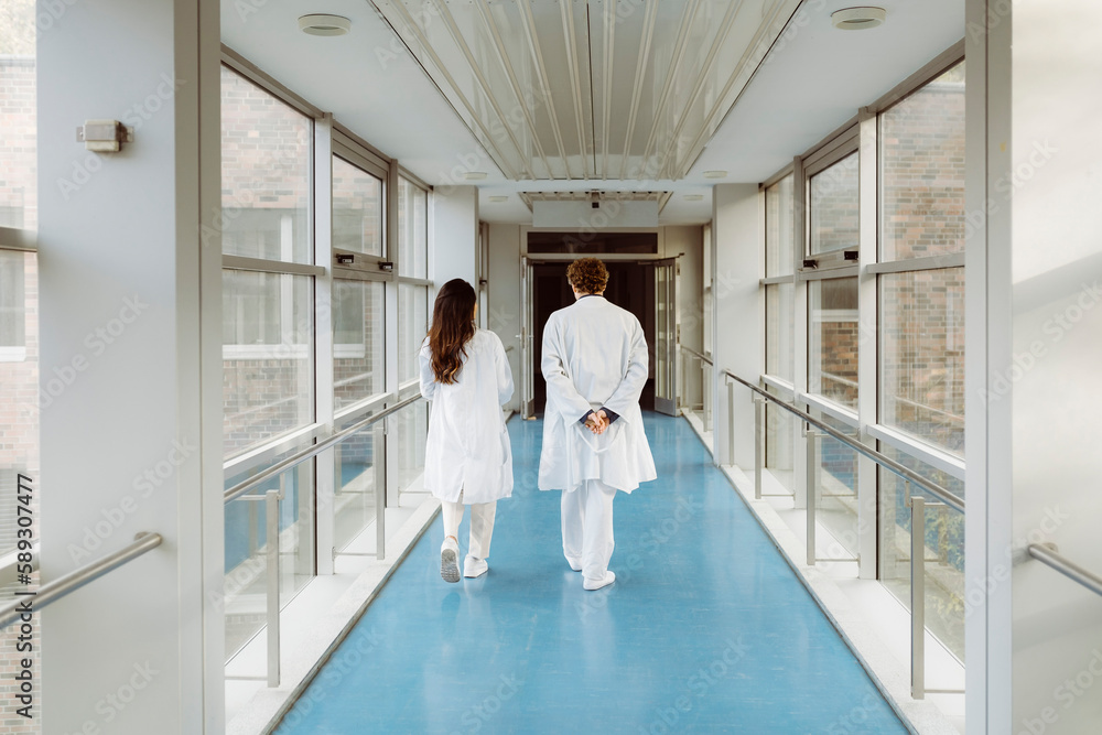 Full length rear view of male and female doctors discussing while walking together in hospital corridor Full length rear view of male and female doctors discussing while walking together in hospital corridor