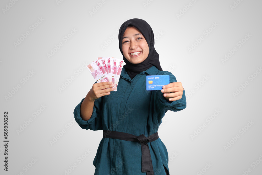 Portrait of cheerful Asian muslim woman with hijab, showing one hundred thousand rupiah while holding a credit card. Financial and savings concept. Isolated image on white background Portrait of cheerful Asian muslim woman with hijab, showing one hundred thousand rupiah while holding a credit card. Financial and savings concept. Isolated image on white background