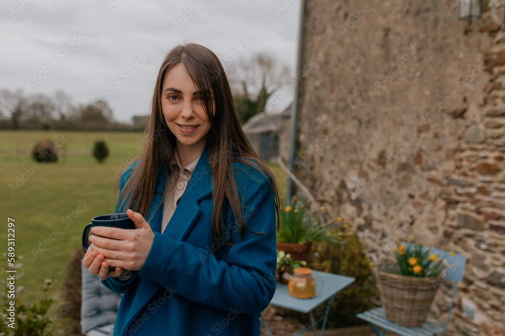 Close up portrait of smiling stylish woman with long hair wearing blue coat is holding coffee while resting at home outdoor in cloudy spring day. 