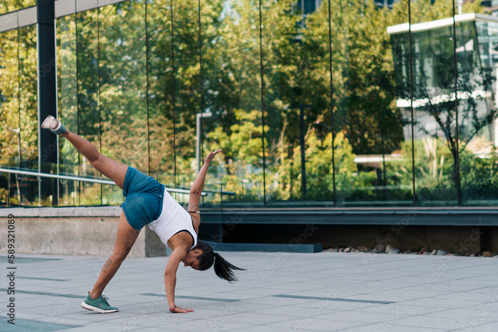 mujer morena con ropa urbana practicando capoeira en la ciudad y haciendo flexiones. - obrazy, fototapety, plakaty 