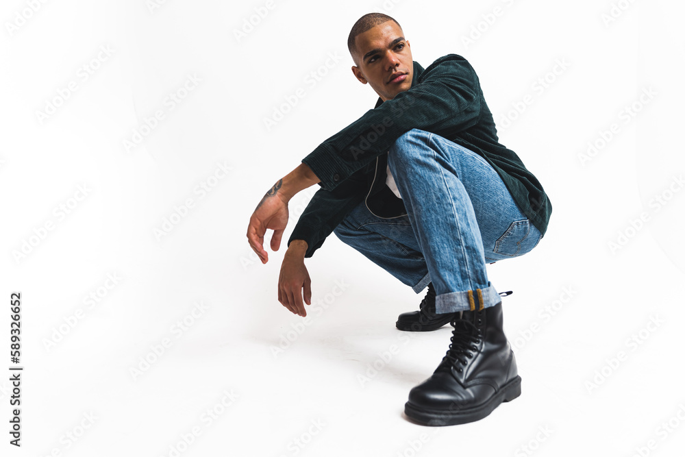 Fototapeta premium Modelling concept. Assured focused young adult guy in fashionable clothes posing on the ground for fashion magazine. White background. High quality photo