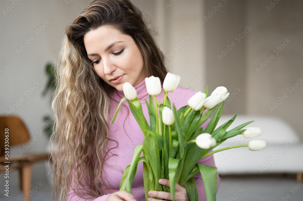 Spring portrait of young curly woman with white tulips in home interior. Woman with a bouquet of flowers, copy space