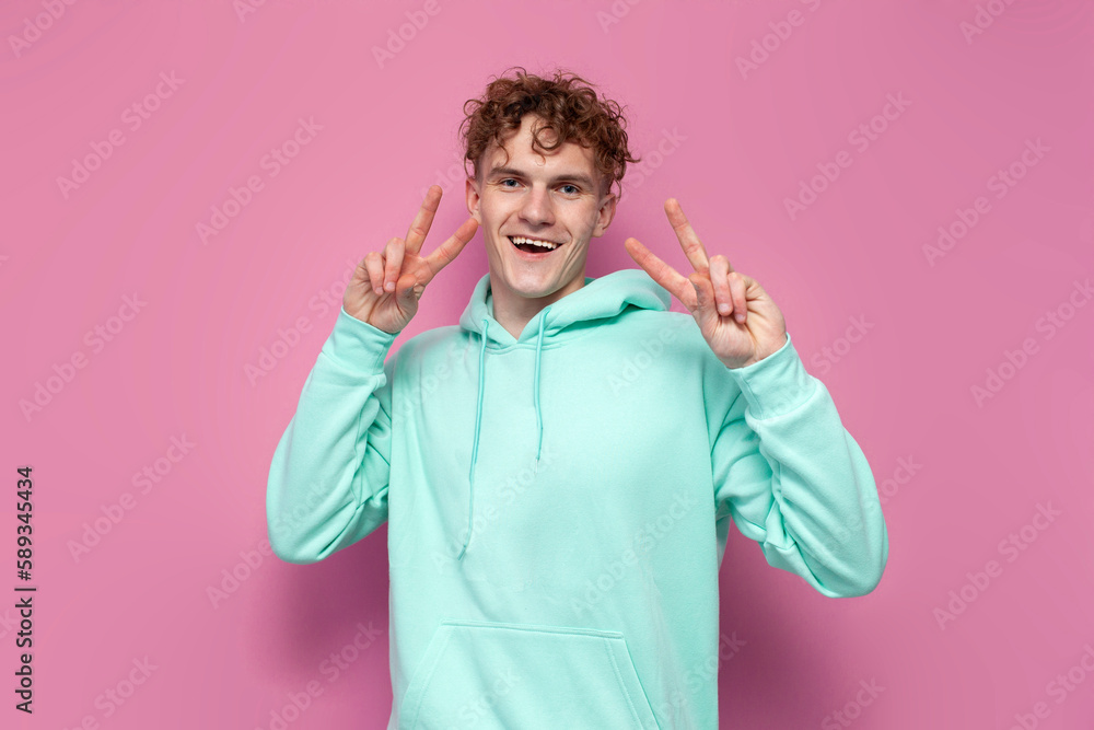 young curly guy in mint hoodie shows gesture of peace with his hands on pink isolated background young curly guy in mint hoodie shows gesture of peace with his hands on pink isolated background