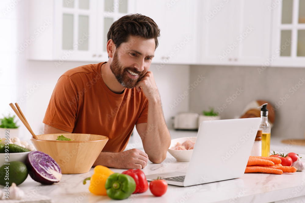Man making dinner while watching online cooking course via laptop in kitchen Man making dinner while watching online cooking course via laptop in kitchen