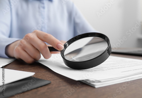 Woman looking at document through magnifier at wooden table indoors, closeup. Searching concept