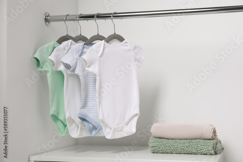 Hangers with baby bodysuits and stack of clothes near white wall. Space for text
