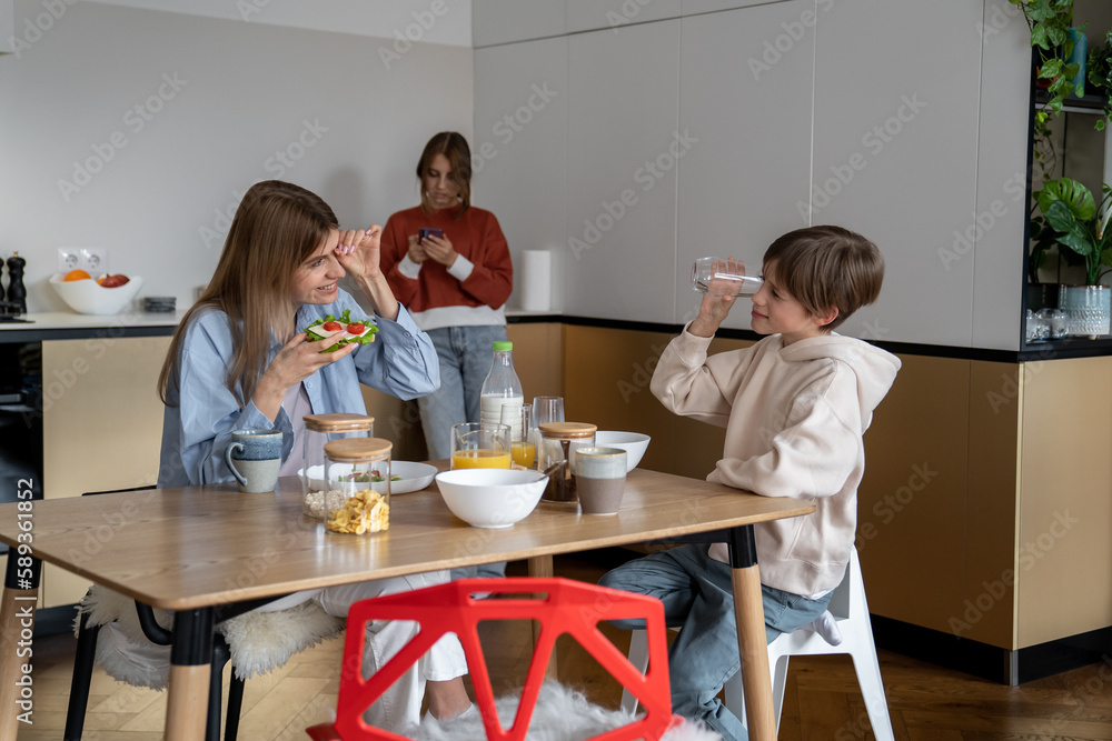 Happy mother and son eating together in morning. Teen girl using smartphone while standing away of family having breakfast without her. Cell phone addiction and family relationships Happy mother and son eating together in morning. Teen girl using smartphone while standing away of family having breakfast without her. Cell phone addiction and family relationships