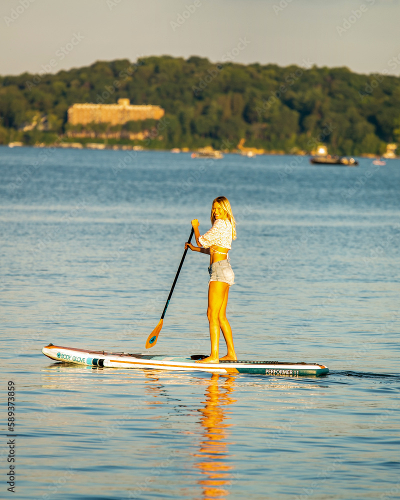 Young blonde woman paddle boarding on the lake wearing shorts and a cropped top smiling, summertime Young blonde woman paddle boarding on the lake wearing shorts and a cropped top smiling, summertime