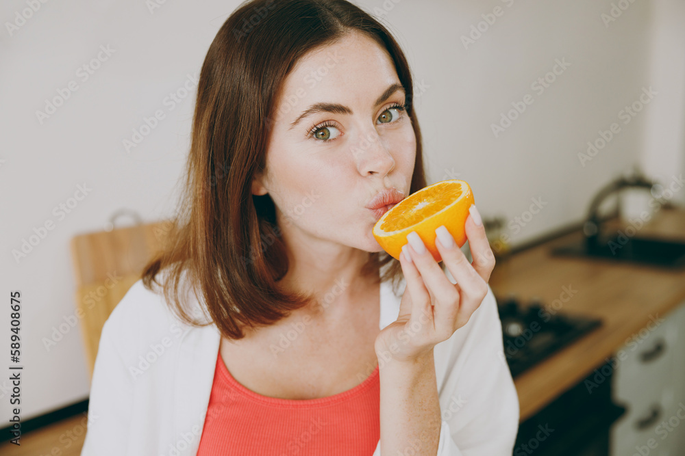 Side view vegetarian fun happy caucasian young housewife woman wear casual clothes biting eat half of orange look camera cooking food breakfast in light kitchen at home alone. Healthy diet concept. Side view vegetarian fun happy caucasian young housewife woman wear casual clothes biting eat half of orange look camera cooking food breakfast in light kitchen at home alone. Healthy diet concept.
