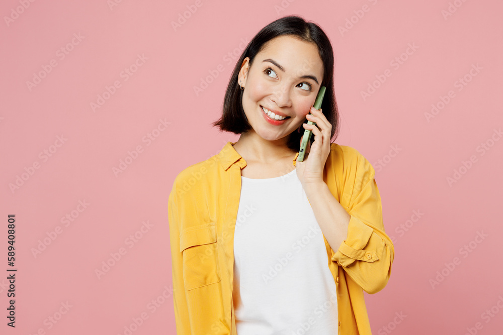 Young woman of Asian ethnicity wear yellow shirt white t-shirt talk speak on mobile cell phone conducting pleasant conversation isolated on plain pastel light pink background studio Lifestyle concept. Young woman of Asian ethnicity wear yellow shirt white t-shirt talk speak on mobile cell phone conducting pleasant conversation isolated on plain pastel light pink background studio Lifestyle concept.