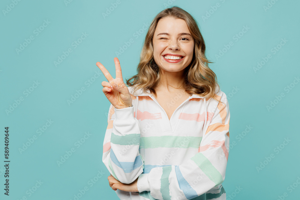 Young smiling cherful fun caucasian blonde woman wearing hoody showing victory sign wink blink eye look camera isolated on plain pastel light blue cyan background studio portrait. Lifestyle concept. Young smiling cherful fun caucasian blonde woman wearing hoody showing victory sign wink blink eye look camera isolated on plain pastel light blue cyan background studio portrait. Lifestyle concept.