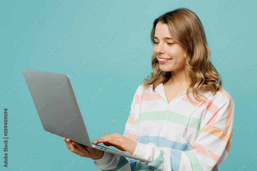 Naklejka premium Side view young IT woman wear striped hoody hold use work on laptop pc computer chatting online browsing internet isolated on plain pastel light blue cyan background studio portrait Lifestyle concept