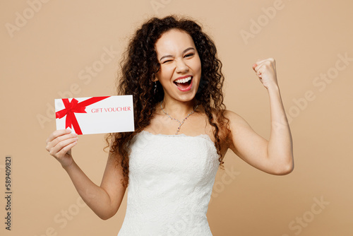 Happy young woman bride in wedding dress posing hold store gift certificate coupon voucher card do winner gesture isolated on plain beige background studio portrait Ceremony celebration party concept
