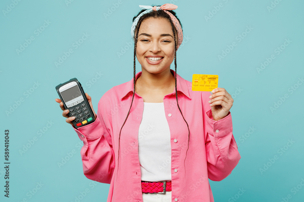Young woman of African American ethnicity in pink shirt white t-shirt headscarf hold wireless bank payment terminal to process acquire credit card isolated on plain pastel light blue cyan background