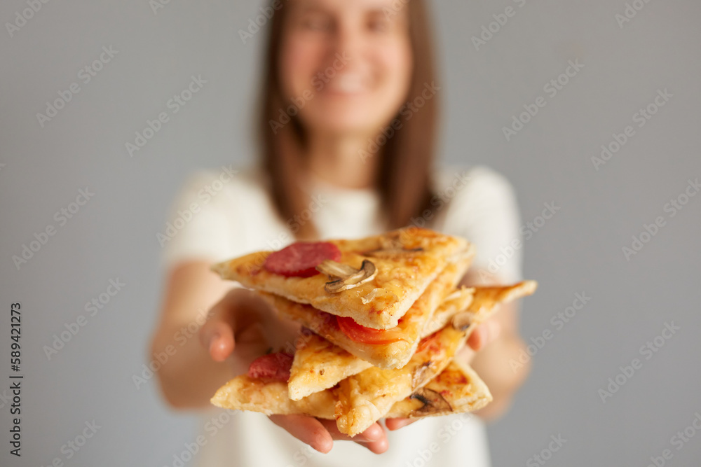 Photo of satisfied woman holding slice of pizza, feels pleased in pizzeria, looks happily directly at camera, offering to eat junk food wearing casual outfit isolated over gray background. Photo of satisfied woman holding slice of pizza, feels pleased in pizzeria, looks happily directly at camera, offering to eat junk food wearing casual outfit isolated over gray background.