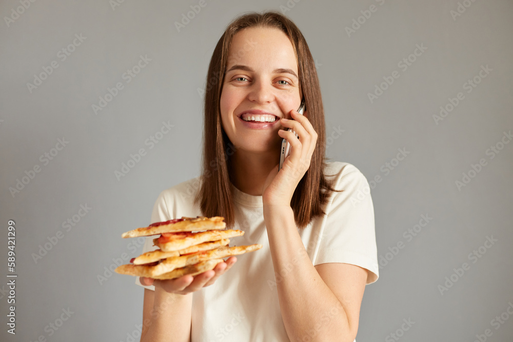 Indoor shot of smiling woman with food isolated over gray background holding slice of pizza and phone, talking with delivery service, ordering supplement. Indoor shot of smiling woman with food isolated over gray background holding slice of pizza and phone, talking with delivery service, ordering supplement.