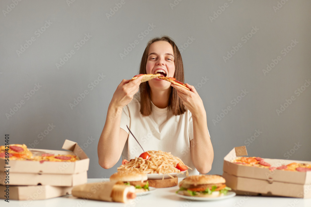 Portrait of extremely hungry woman with brown hair wearing white T-shirt sitting at table isolated over gray background, biting two slices of pizza same time. Portrait of extremely hungry woman with brown hair wearing white T-shirt sitting at table isolated over gray background, biting two slices of pizza same time.