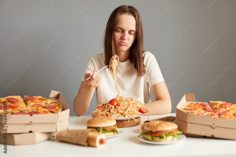 Indoor shot of frustrated sad woman with brown hair wearing white T-shirt sitting at table isolated over gray background, eating pasta, being disappointed, being not hungry. Indoor shot of frustrated sad woman with brown hair wearing white T-shirt sitting at table isolated over gray background, eating pasta, being disappointed, being not hungry.