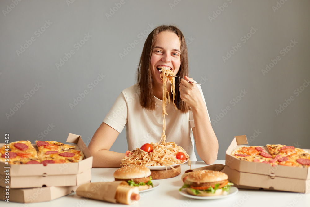 Portrait of smiling charming woman with brown hair wearing white T-shirt sitting at table isolated over gray background, eating pasta, having delicious dinner. Portrait of smiling charming woman with brown hair wearing white T-shirt sitting at table isolated over gray background, eating pasta, having delicious dinner.