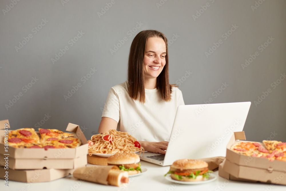 Indoor shot of smiling woman working on laptop computer, typing, communicating online, sitting at table surrounded with fast food isolated over gray background. Indoor shot of smiling woman working on laptop computer, typing, communicating online, sitting at table surrounded with fast food isolated over gray background.