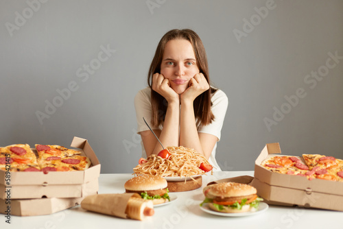 Portrait of sad upset bored woman with brown hair wearing white T-shirt sitting at table among junk food wants healthy dish isolated over gray background  looking at camera with sorrow.