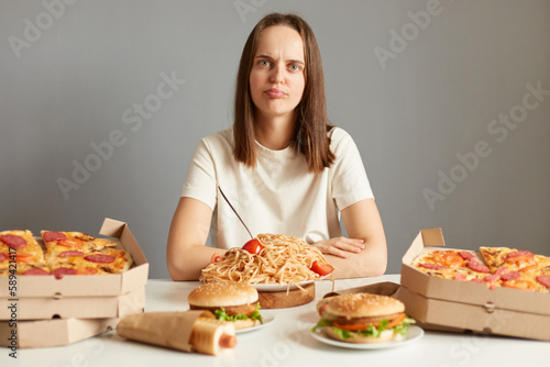 Portrait of upset irritated woman with brown hair wearing white T-shirt sitting at table isolated over gray background, being dissapointed of junk food menu, prefers healthy eating.