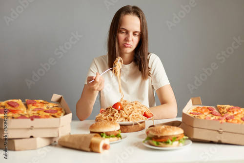 Indoor shot of frustrated sad woman with brown hair wearing white T-shirt sitting at table isolated over gray background  eating pasta  being disappointed  being not hungry.
