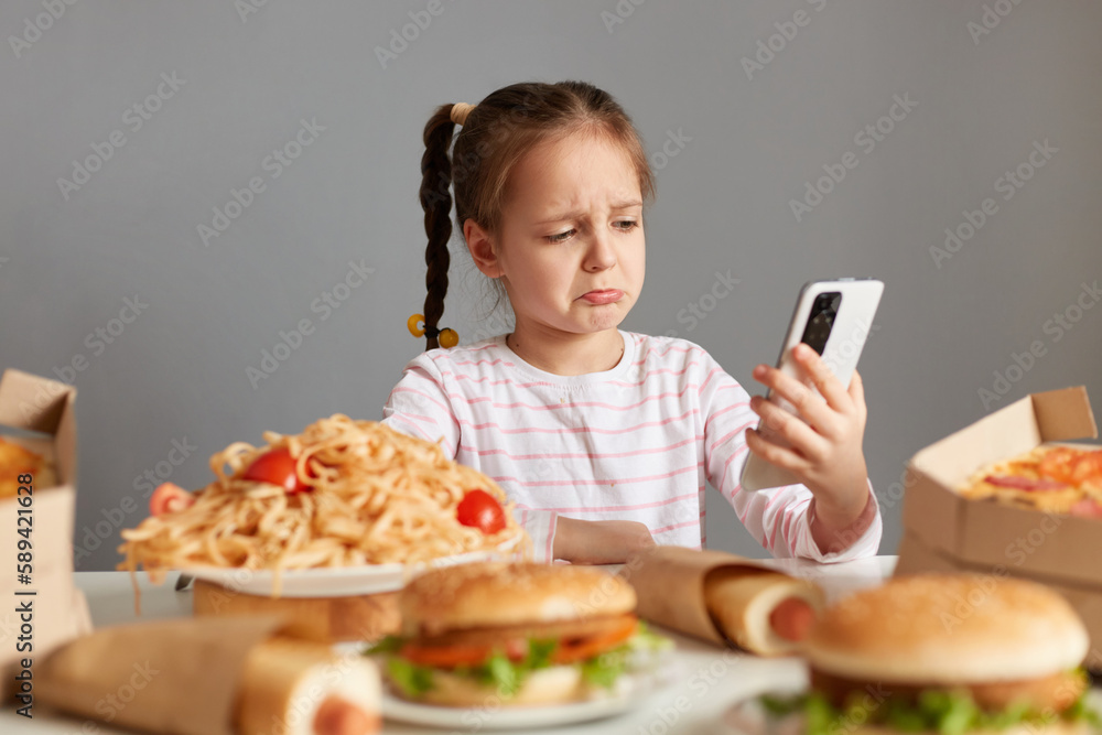 Indoor shot of sad crying little girl with pigtails sitting at table pizza, hot dogs, burgers, talking via video call, keeps diet, can't eat delicious fast food, isolated over gray background