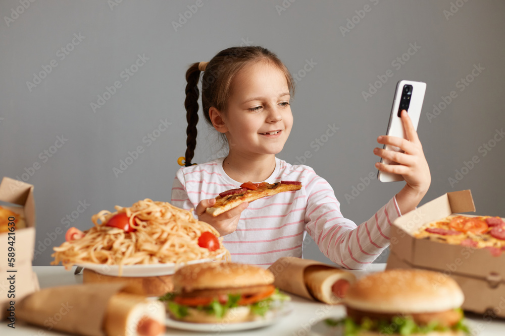 Photo of joyful little girl with pigtails sitting at table isolated over gray background, using mobile phone, having video call, boasting with big variety of unhealthy tasty dishes. Photo of joyful little girl with pigtails sitting at table isolated over gray background, using mobile phone, having video call, boasting with big variety of unhealthy tasty dishes.