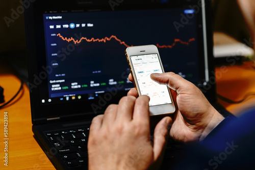 Businessman trader investor analyst using mobile phone app analytics for financial market analysis,phone with graphs and diagrams on screen,using smartphone.sitting at work desk with laptop.