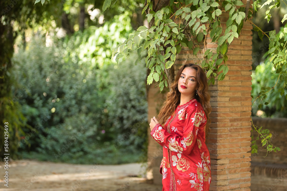 Beautiful young woman in a typical Moroccan red suit, embroidered with gold and silver threads, posing next to a brick column with a vine. Concept beauty, ethnicity, typical suits, Marrakech, Arab. Beautiful young woman in a typical Moroccan red suit, embroidered with gold and silver threads, posing next to a brick column with a vine. Concept beauty, ethnicity, typical suits, Marrakech, Arab.