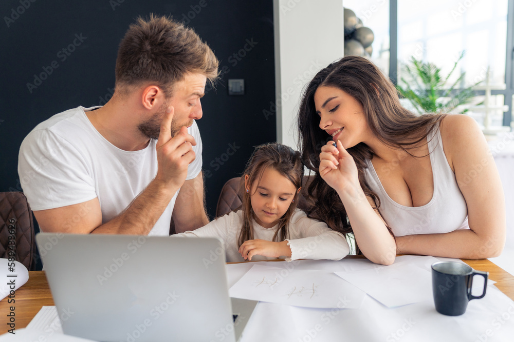 Young parents teach their daughter to write, helping her with her homework, untidy desk and laptop. Concept happy healthy family Young parents teach their daughter to write, helping her with her homework, untidy desk and laptop. Concept happy healthy family