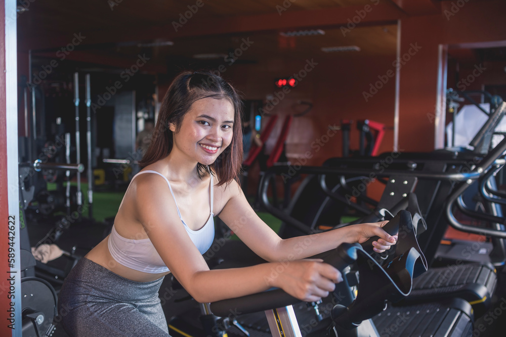 A fit asian woman smiles while doing cardio on a stationary bike. Sweating out and burning calories at the gym. A fit asian woman smiles while doing cardio on a stationary bike. Sweating out and burning calories at the gym.