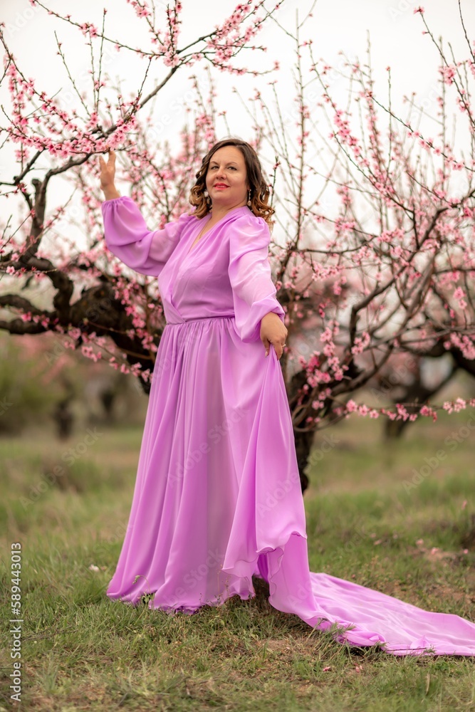 Woman peach blossom. Happy curly woman in pink dress walking in the garden of blossoming peach trees in spring Woman peach blossom. Happy curly woman in pink dress walking in the garden of blossoming peach trees in spring