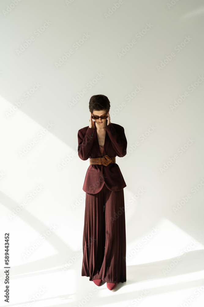 full length of brunette model in stylish jacket and palazzo pants adjusting sunglasses on grey background with lighting. full length of brunette model in stylish jacket and palazzo pants adjusting sunglasses on grey background with lighting.