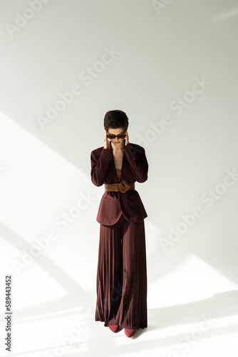 full length of brunette model in stylish jacket and palazzo pants adjusting sunglasses on grey background with lighting.