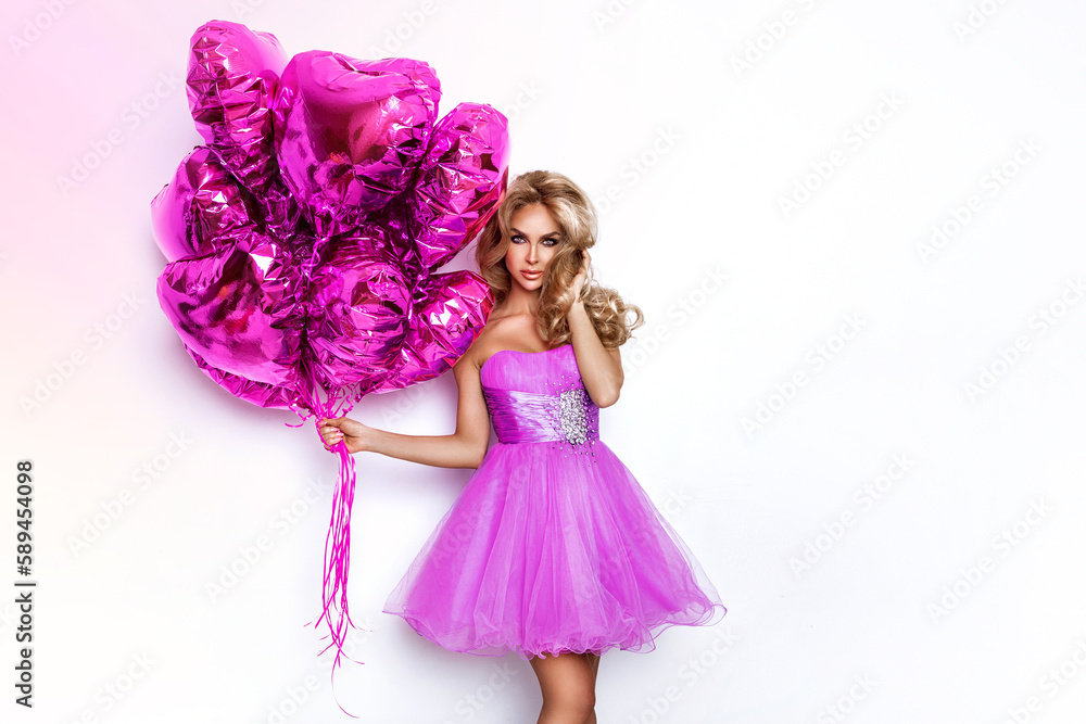 Sexy blonde woman in lilac summer dress is holding pink balloons and is posing on white background in studio.