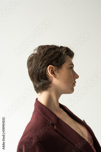 profile of stylish brunette woman in cuff earring and blazer isolated on grey.