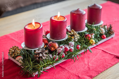 Brennende rote Adventskerzen mit Dekoration auf einem Holztisch im Wohnzimmer auf rotem Stoffstuch zur Weihnachtszeit, Deutschland