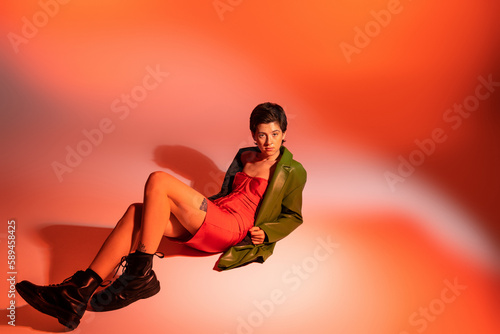 full length of tattooed woman in red corset dress and green jacket with black boots lying on colorful background.