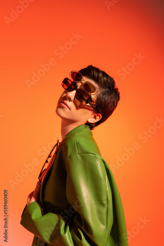 stylish brunette woman posing in several sunglasses and green leather jacket on orange background.