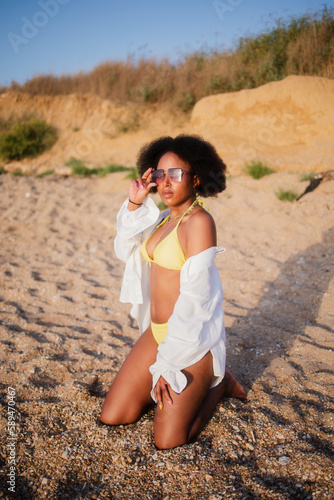 African american woman in a yellow swimsuit and a white shirt in a white shirt sits on the beach relaxing. Concept: relaxation, tour operator, ocean, hotel, beach, vacation