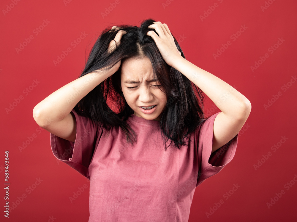 A portrait of a young Indonesian (Asian) woman wearing a pink shirt, looking dizzy and holding her head with both hands. Isolated with a red background A portrait of a young Indonesian (Asian) woman wearing a pink shirt, looking dizzy and holding her head with both hands. Isolated with a red background