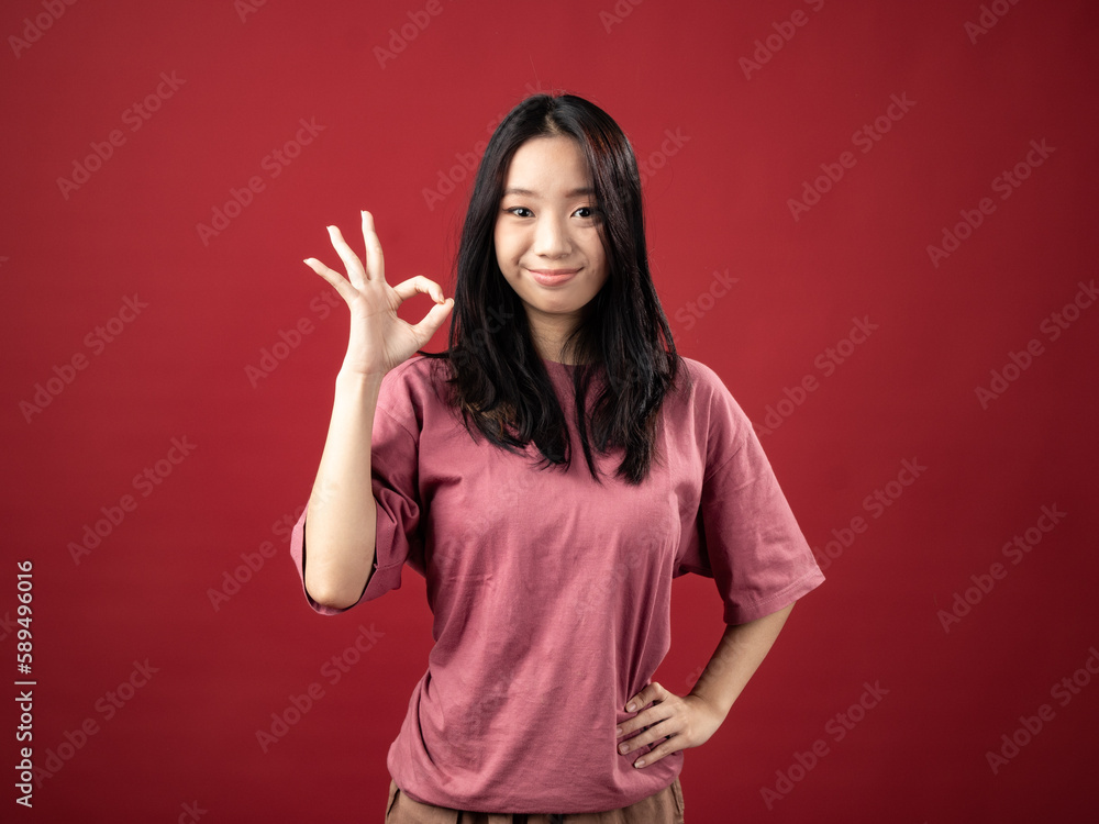 A studio portrait of a young Indonesian (Asian) woman wearing a pink shirt and displaying the OK sign with her hand. Isolated with a red background. A studio portrait of a young Indonesian (Asian) woman wearing a pink shirt and displaying the OK sign with her hand. Isolated with a red background.