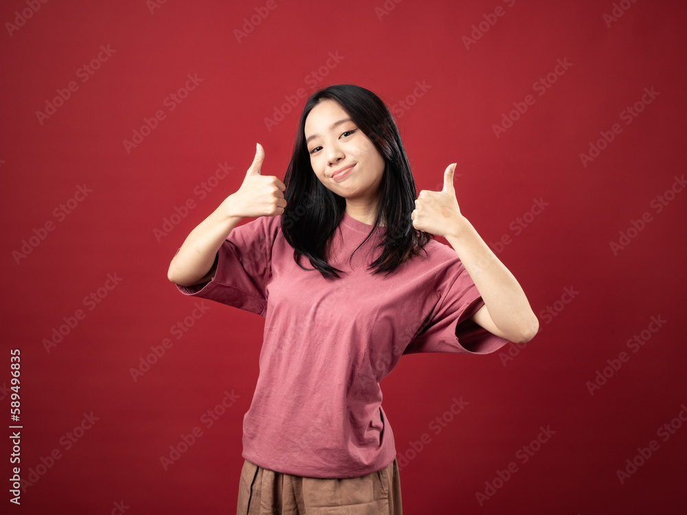 A studio portrait of a young Indonesian (Asian) woman wearing a pink shirt and pointing with her thumb. Isolated with a red background. A studio portrait of a young Indonesian (Asian) woman wearing a pink shirt and pointing with her thumb. Isolated with a red background.