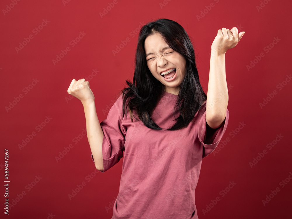 A studio portrait of a young Indonesian woman (Asian) wearing a pink shirt, seen celebrating and raising both hands. Isolated with a red background A studio portrait of a young Indonesian woman (Asian) wearing a pink shirt, seen celebrating and raising both hands. Isolated with a red background
