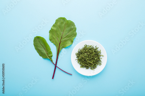 Fresh green beetroot leaves and dry chopped leaves on white plate on light blue table background. Pastel color. Spice preparation and storage. Closeup. Top down view.